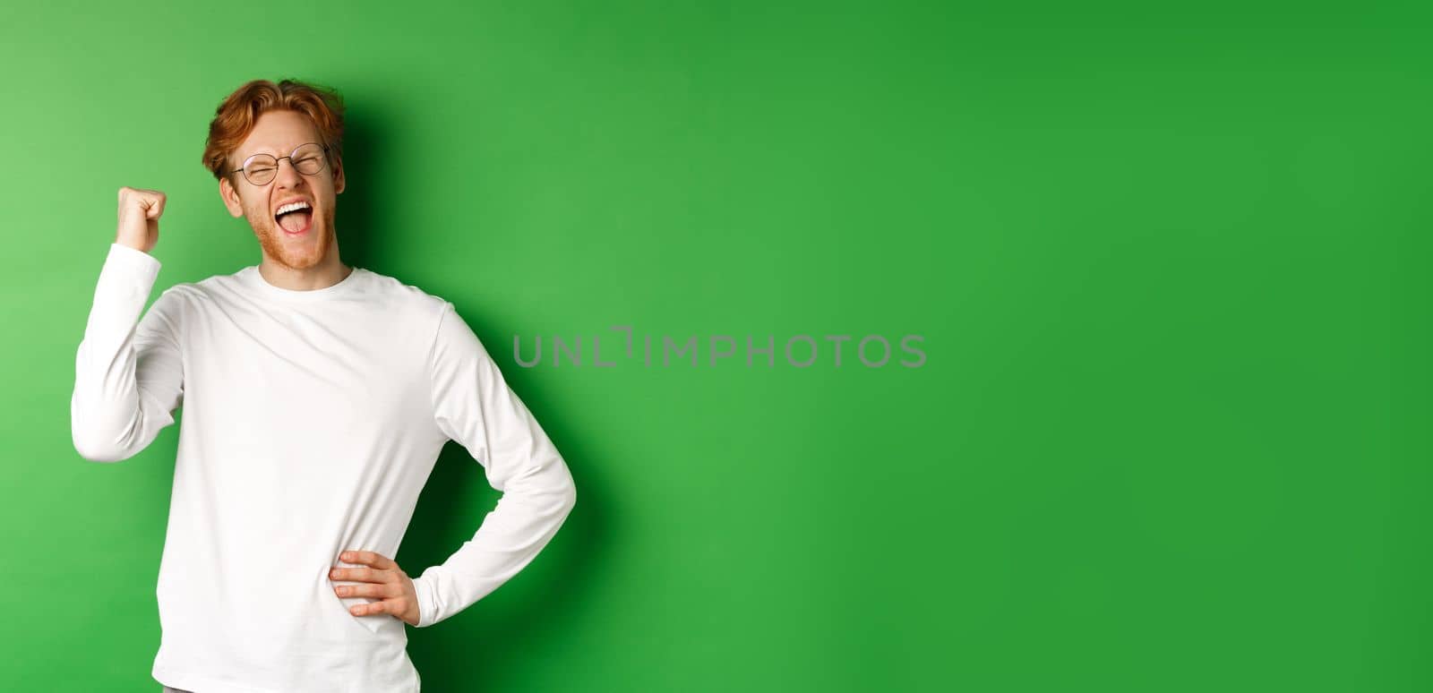 Young cheerful guy scream of joy, winning prize and celebrating, making fist pump gesture and smiling happy, standing over green background by Benzoix