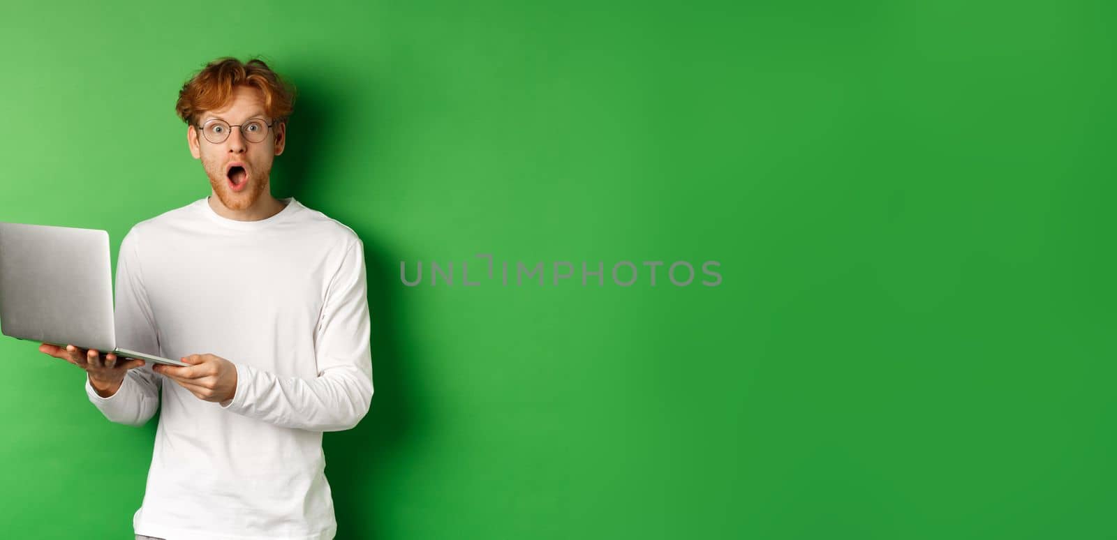 Impressed young man in glasses drop jaw and staring amazed after reading promo on laptop, standing over green background by Benzoix