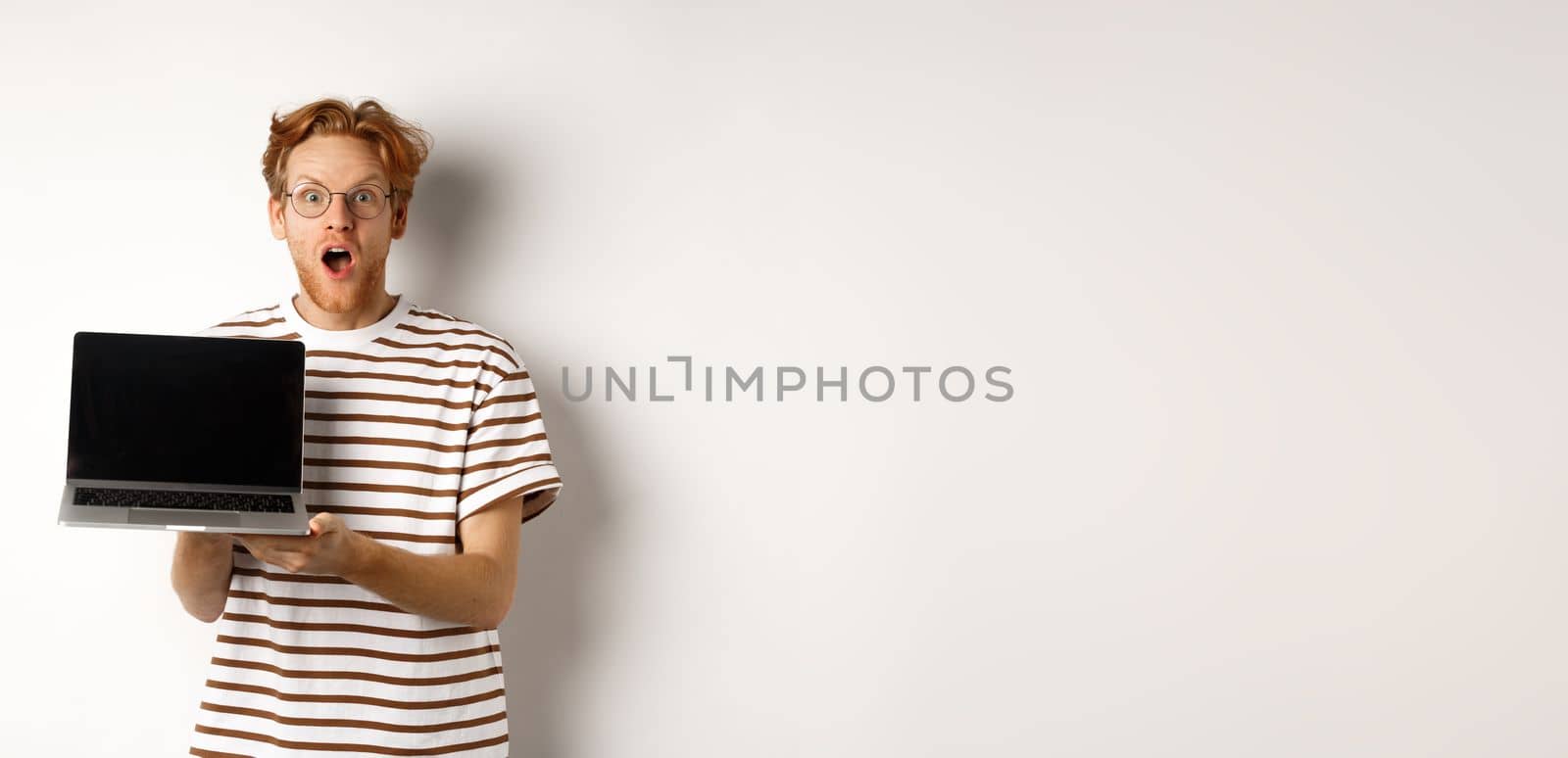 Excited redhead man drop jaw, showing laptop screen logo or website, demonstrate promo offer on display, standing against white background by Benzoix