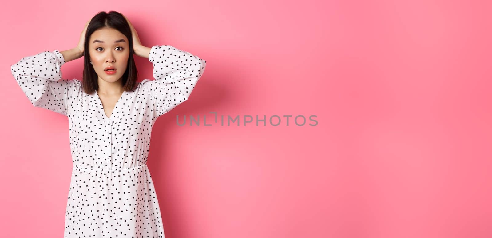 Shocked and troubled asian woman looking confused, holding hands on head, having problem, standing over pink background by Benzoix
