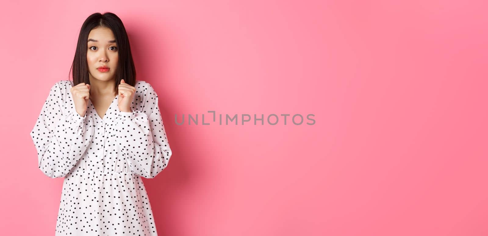 Scared cute asian girl in dress looking worried, feeling frightened and staring at camera nervously, standing over pink background.