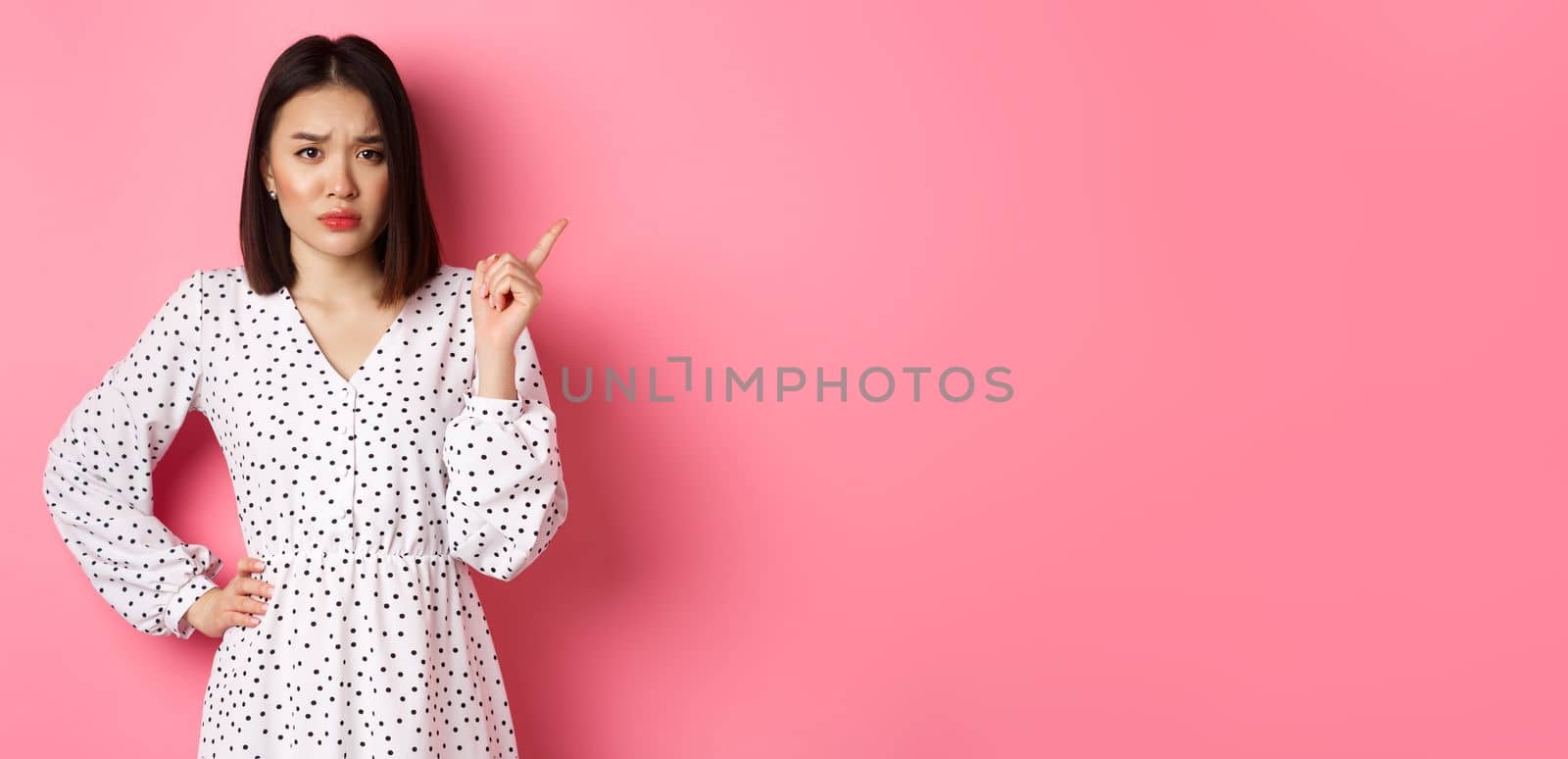 Sad and gloomy asian girl frowning, pointing finger at upper right corner and staring at camera unhappy, standing over pink background by Benzoix