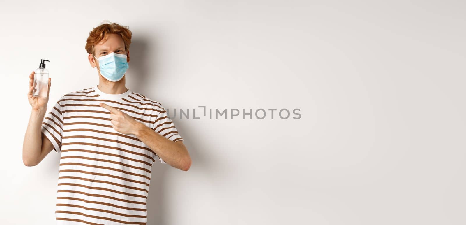 Covid-19, health and lifestyle concept. Cheerful redhead man in face mask pointing finger at hand sanitizer, recommending antiseptic, white background by Benzoix