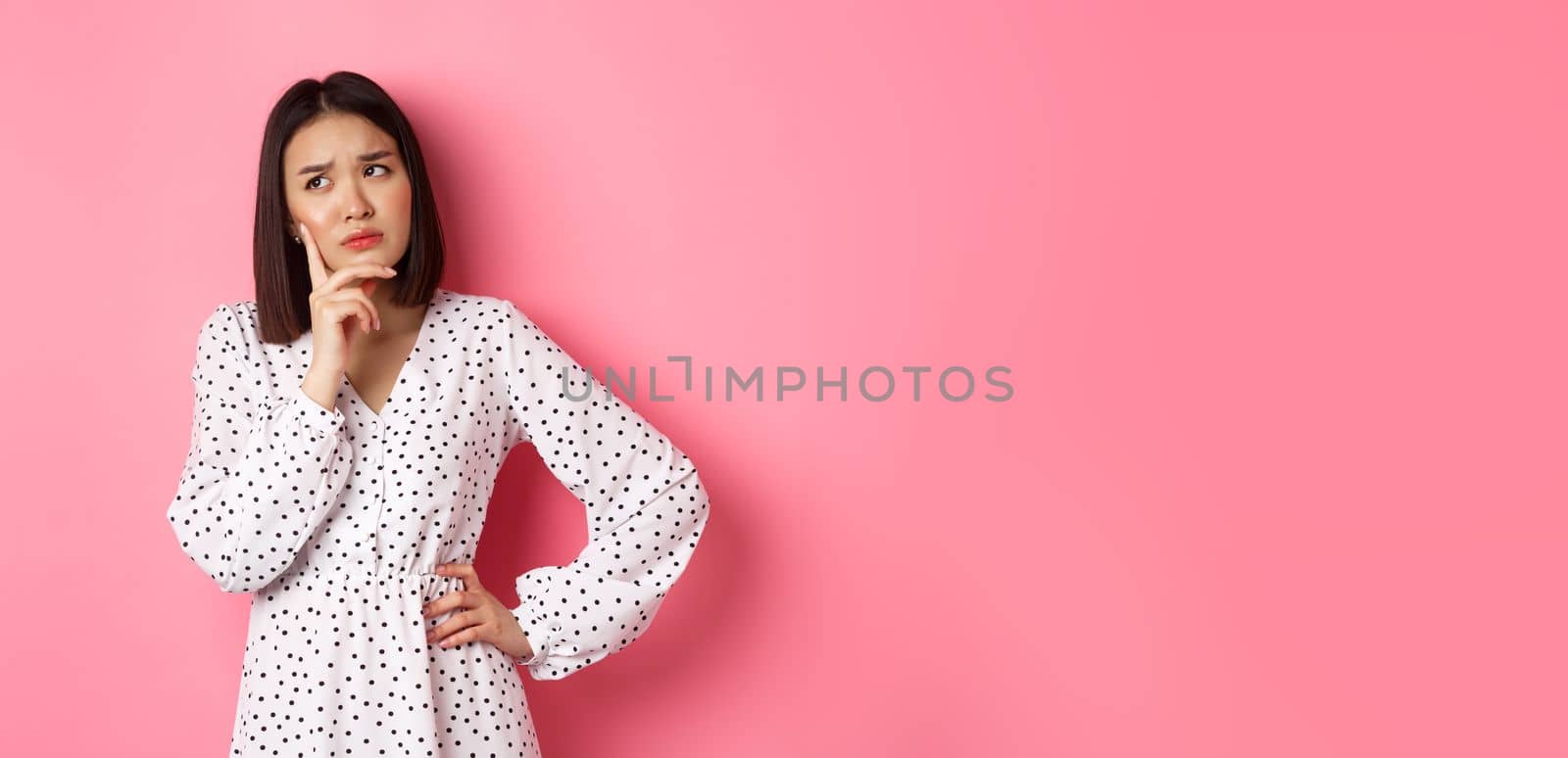 Concerned young asian female model making decision, seriously thinking, looking at upper left corner and choosing, standing over pink background.