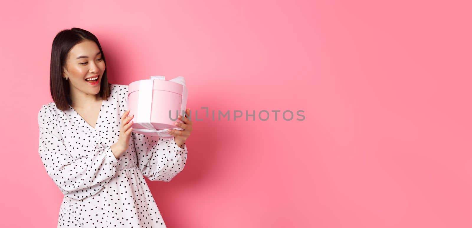 Valentines day, celebration concept. Beautiful asian woman holding romantic gift box, smiling happy, standing over pink background by Benzoix