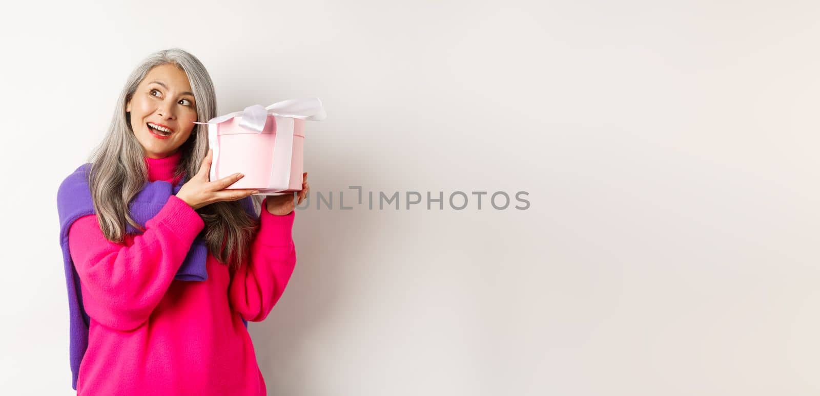 Holidays and valentines day concept. Happy asian mature woman shaking box with gift, guessing what inside present, standing over white background by Benzoix