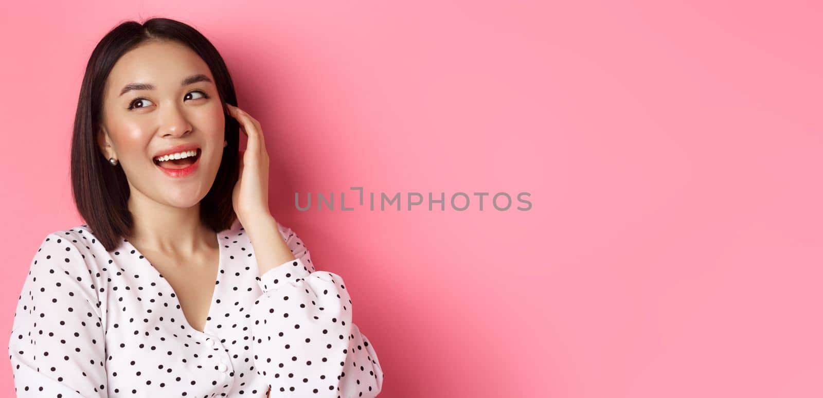 Close-up of flirty asian woman sighing, looking dreamy left and smiling, standing over pink background by Benzoix