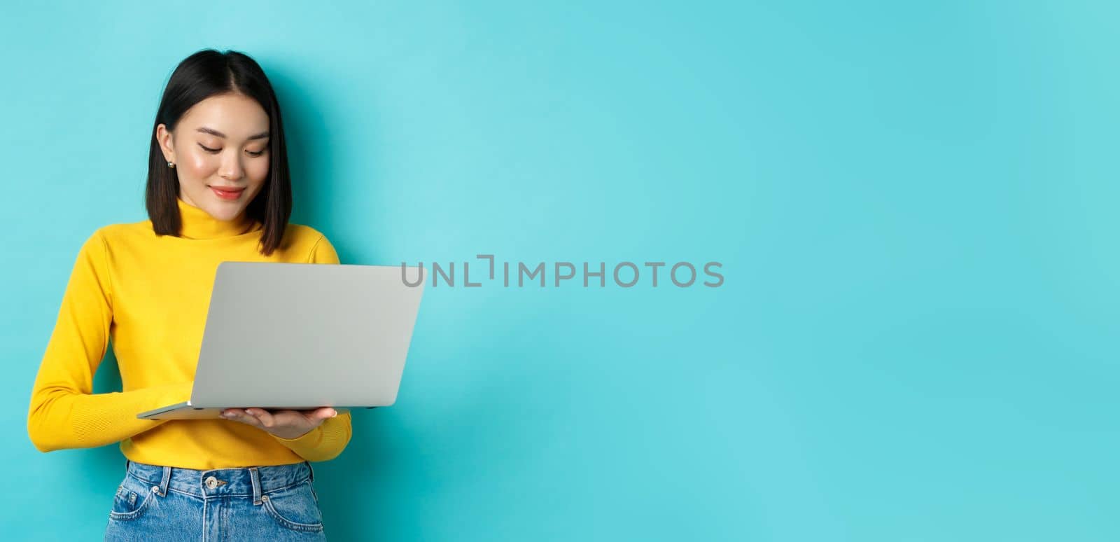 Beautiful asian female student working on laptop, typing on keyboard and looking at screen with pleased smile, standing over blue background.