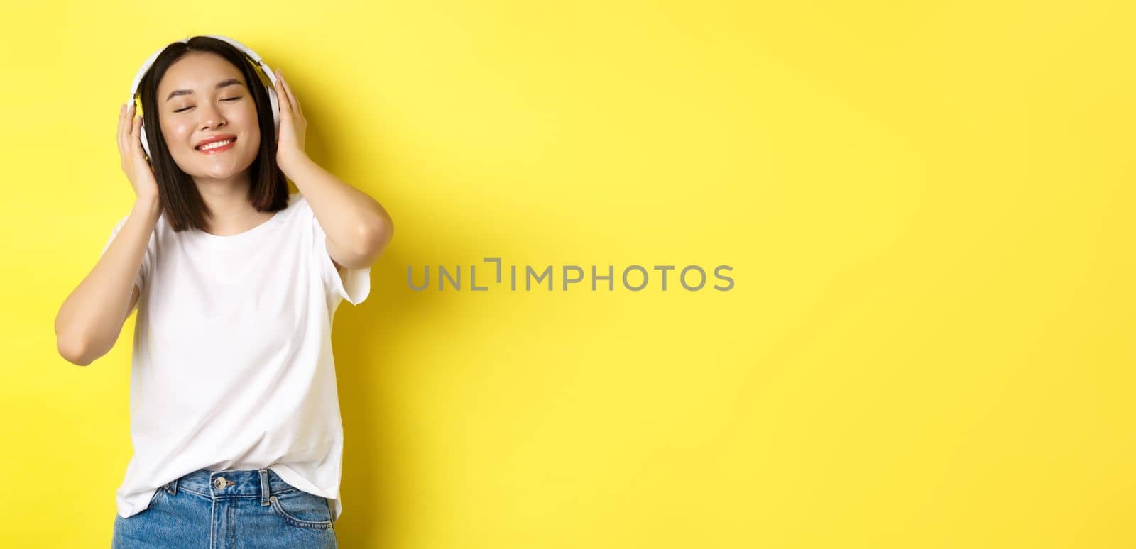 Attractive asian woman enjoying listening music in wireless headphones, smiling pleased and dancing with eyes closed, standing over yellow background.