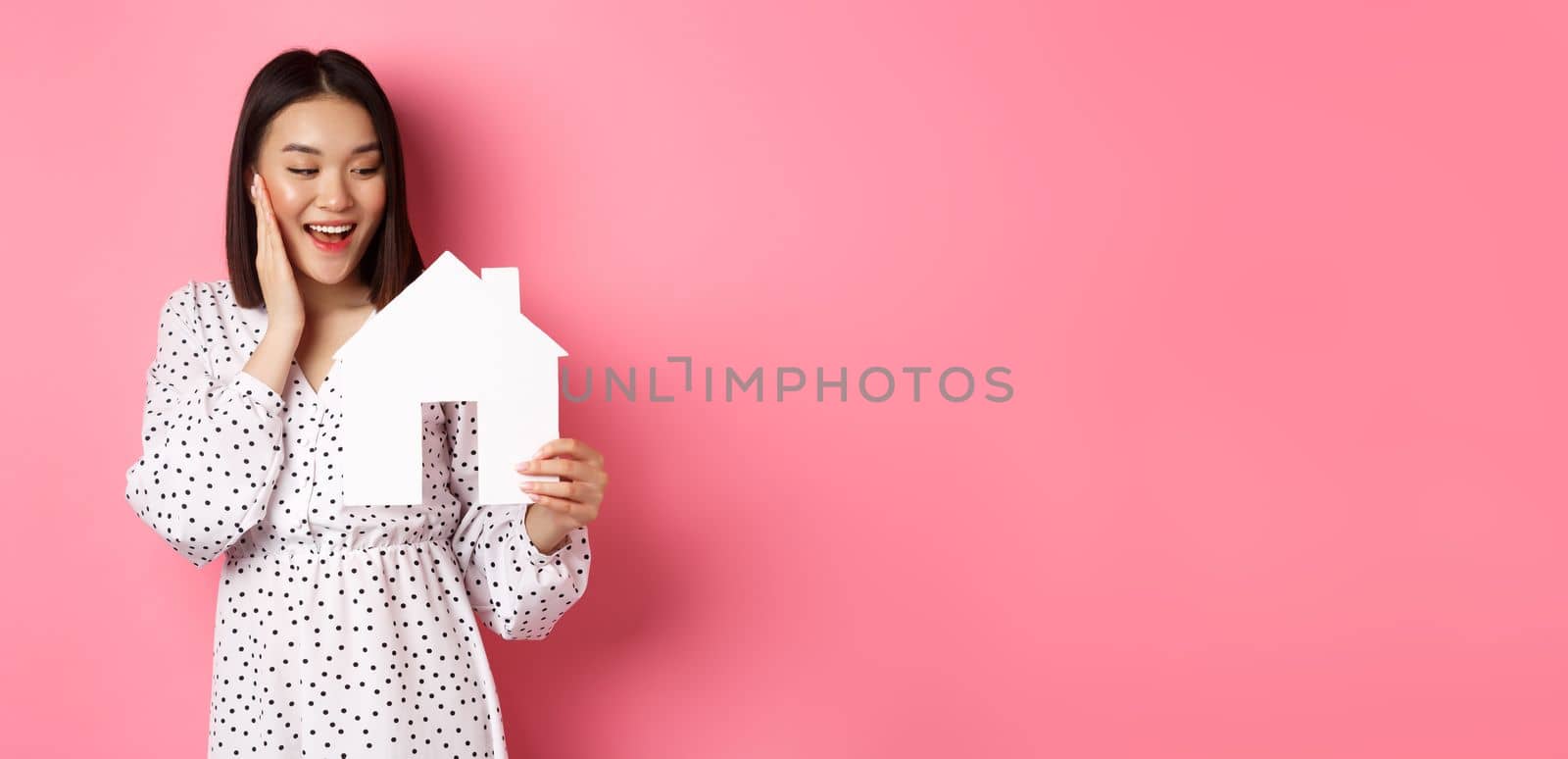 Real estate. Cute asian woman searching for apartment, looking happy at paper house model, standing against pink background by Benzoix