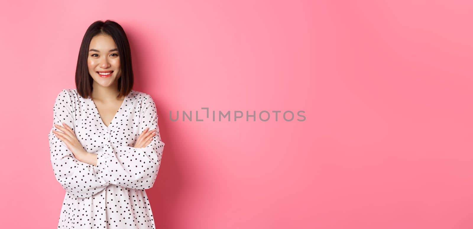 Pretty young asian woman in spring dress smiling, cross arms on chest and looking confident, standing over pink background.