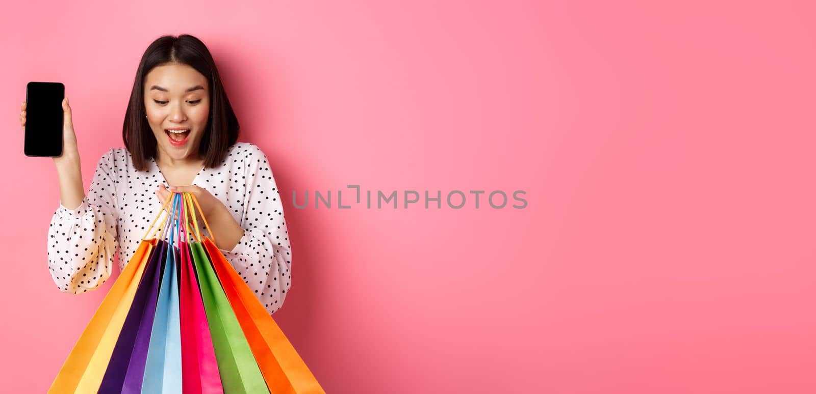 Attractive asian woman showing smartphone app and shopping bags, buying online via application, standing over pink background.