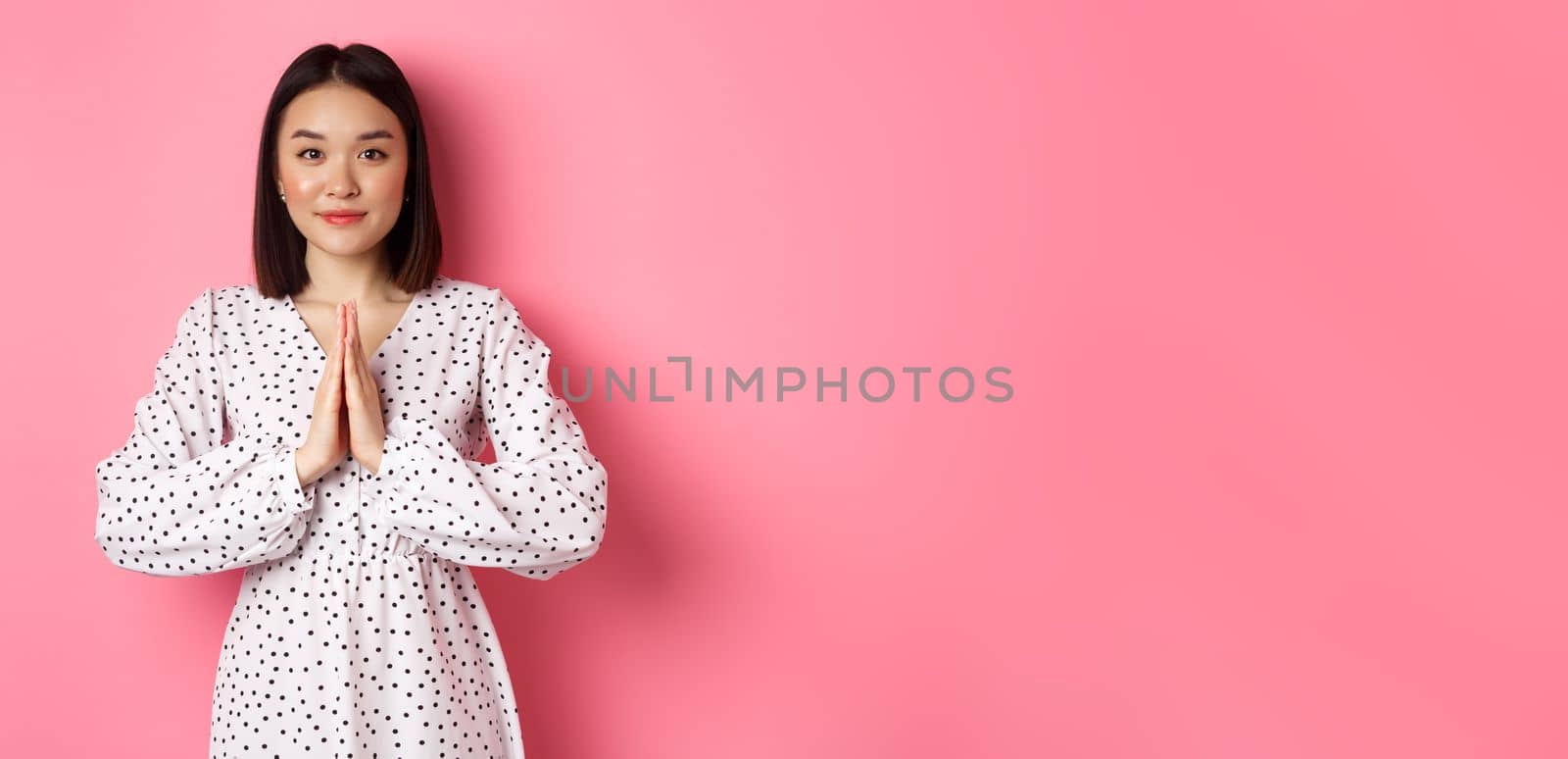 Beautiful asian lady in dress asking for help, holding hands in pray or namaste gesture, thanking you, standing over pink background.