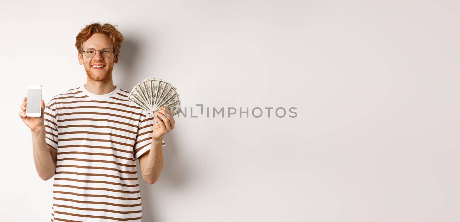 Smiling young redhead man in glasses showing smartphone blank screen and money, standing over white background by Benzoix
