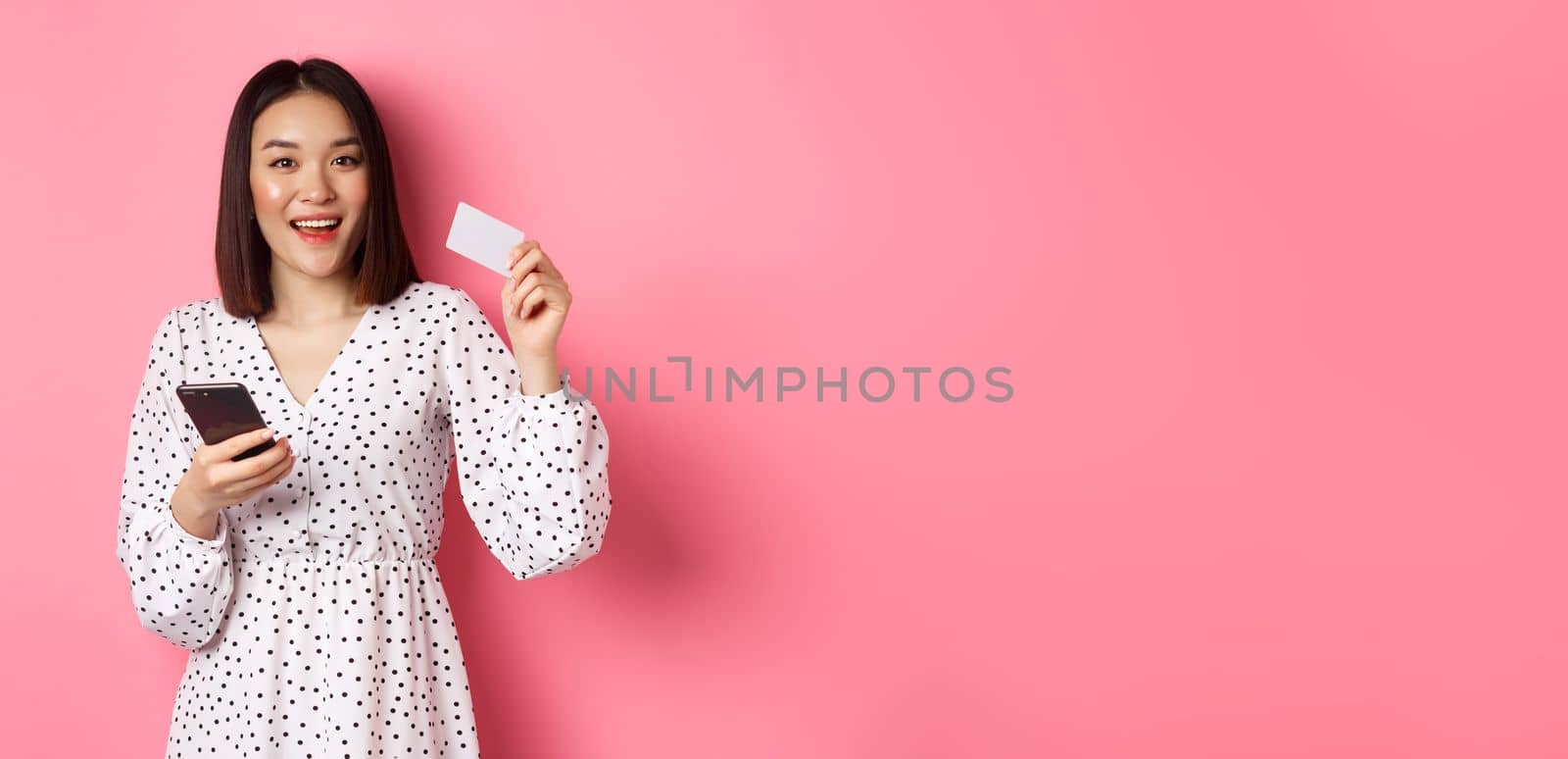 Attractive young asian woman order online, holding credit card and mobile phone, making internet purchase, standing happy over pink background.