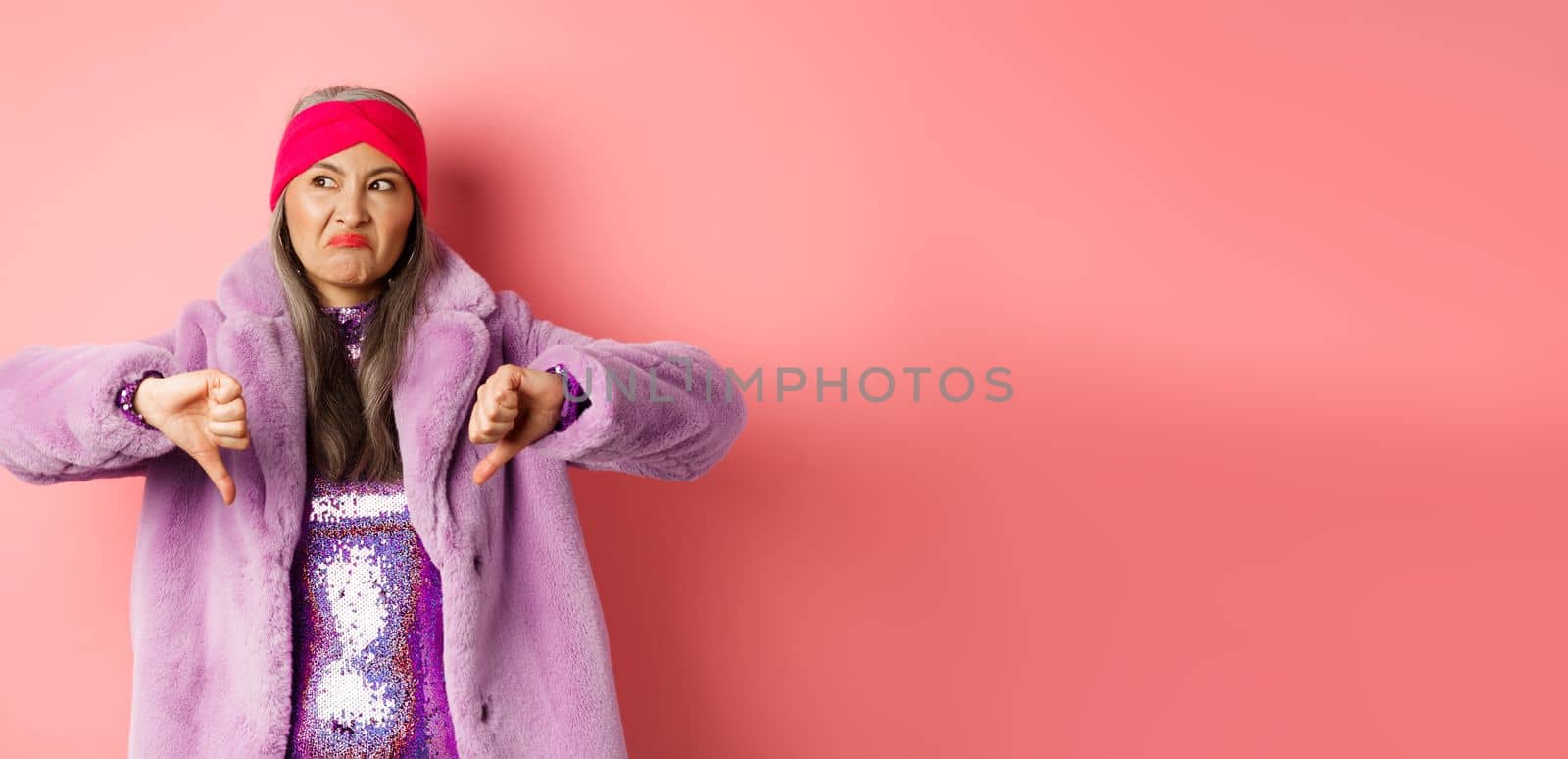 Fashion and shopping concept. Disappointed stylish senior woman showing thumbs-down, looking displeased and judgemental, being dissatisfied, standing over pink background.