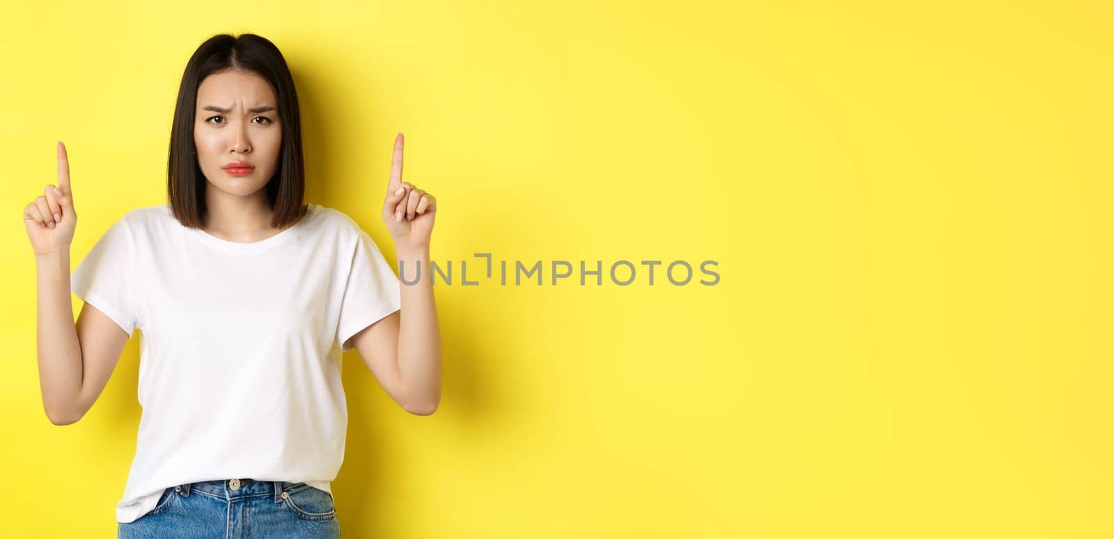 Beauty and fashion concept. Beautiful asian woman in white t-shirt pointing fingers up, standing over yellow background.