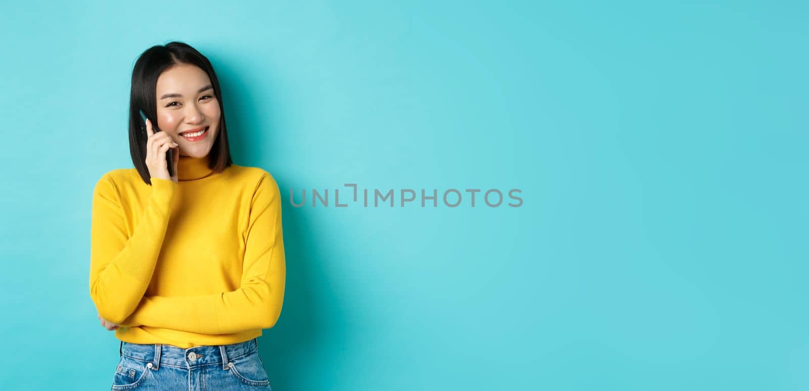 Young stylish asian woman talking on phone, calling friend and smiling, standing with smartphone against blue background.