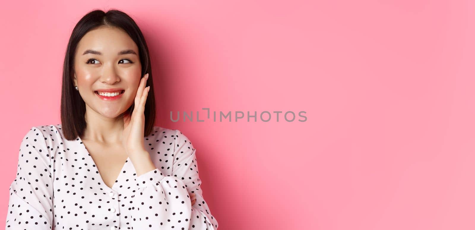 Coquettish asian woman in blushing, touching cheek and looking left amused, standing over pink background by Benzoix