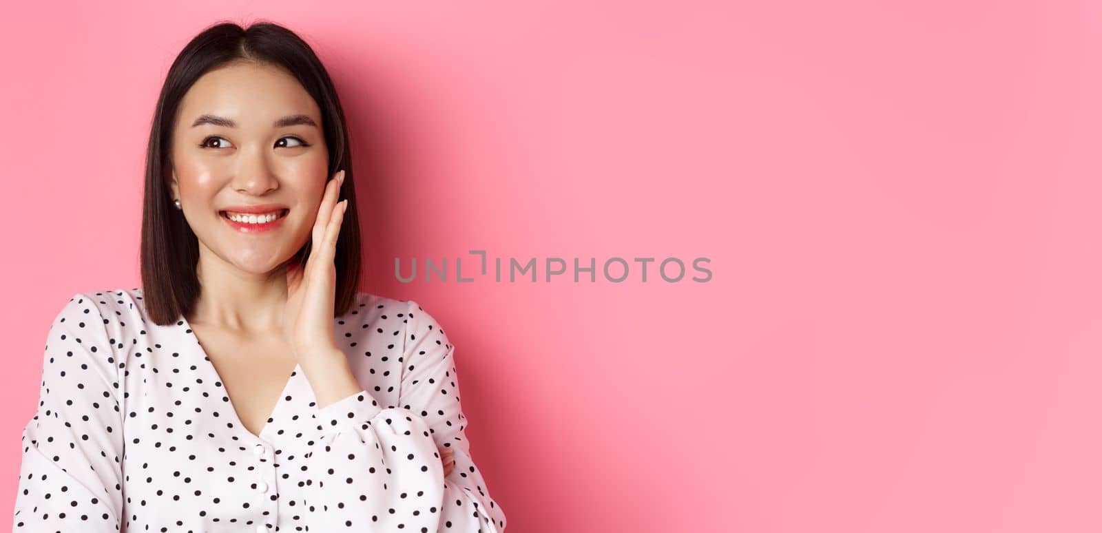 Coquettish asian woman in blushing, touching cheek and looking left amused, standing over pink background.