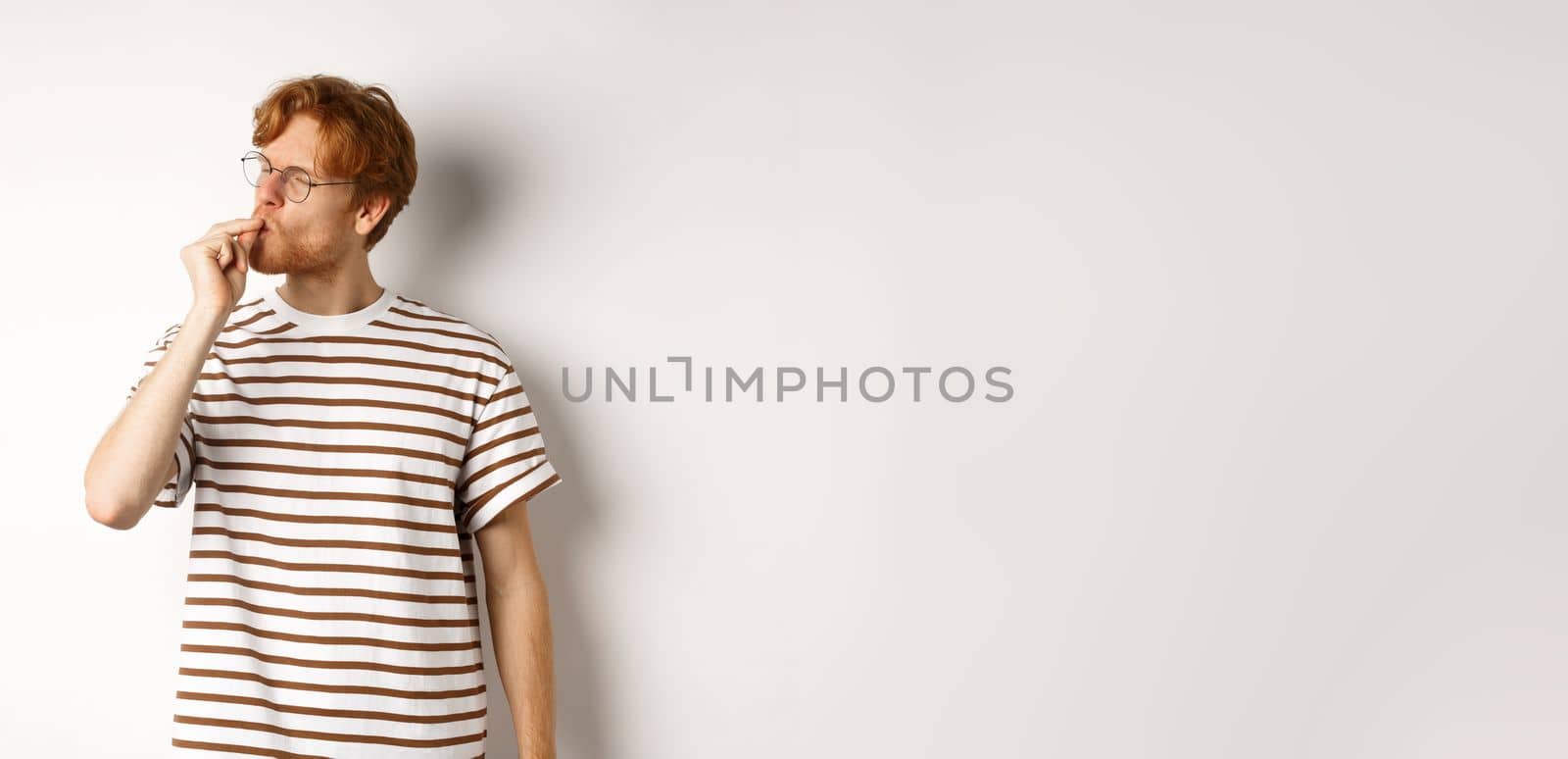 Image of satisfied young man with red hair and glasses kissing fingers, showing chefs kiss gesture to praise something perfect, standing over white background.