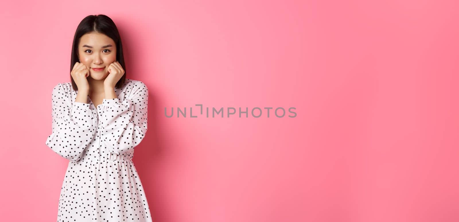 Cute and shy asian girl blushing, touching cheeks and looking at camera silly, standing against pink background.