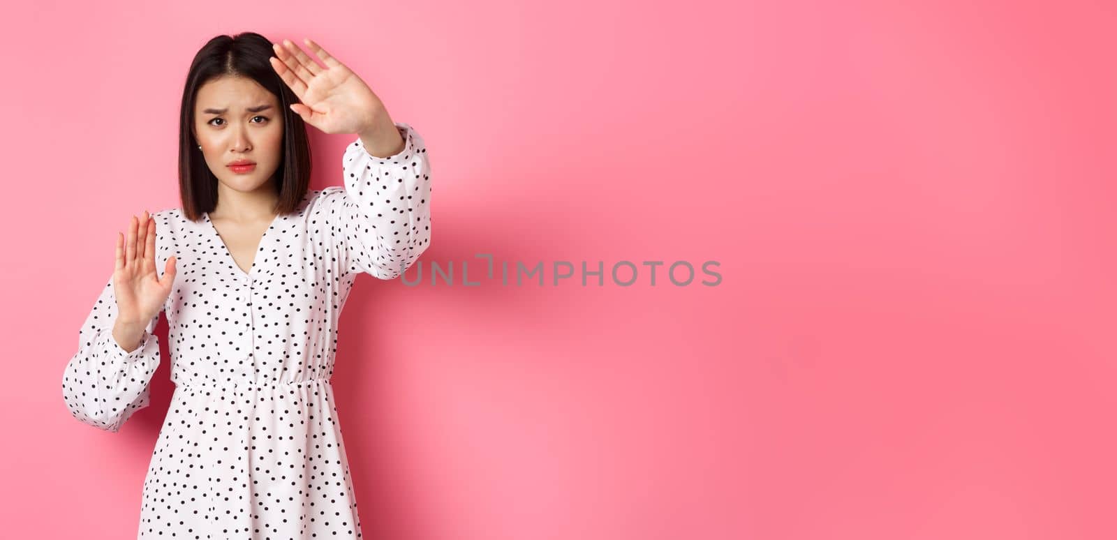 Timid and scared asian woman defending herself, raising arms in protection, victim being attacked, standing over pink background by Benzoix