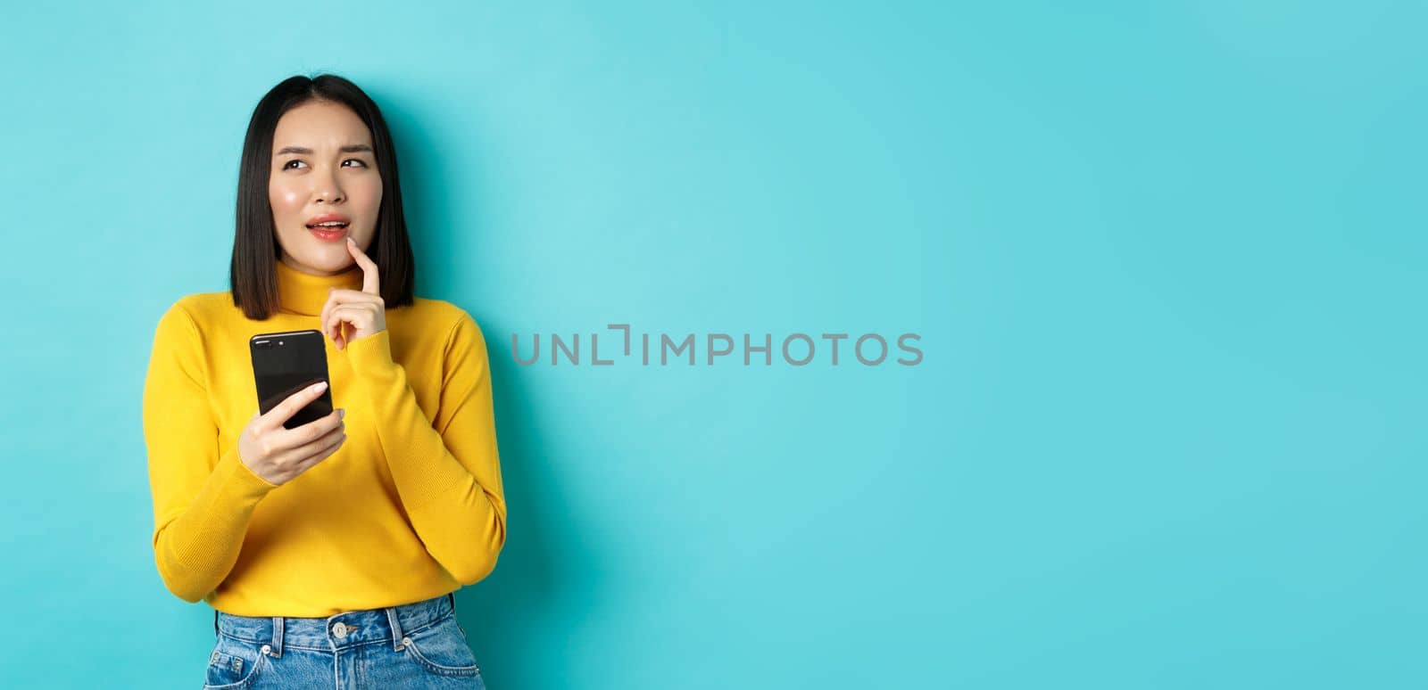 Pensive asian girl holding smartphone and thinking what to order online, standing over blue background.