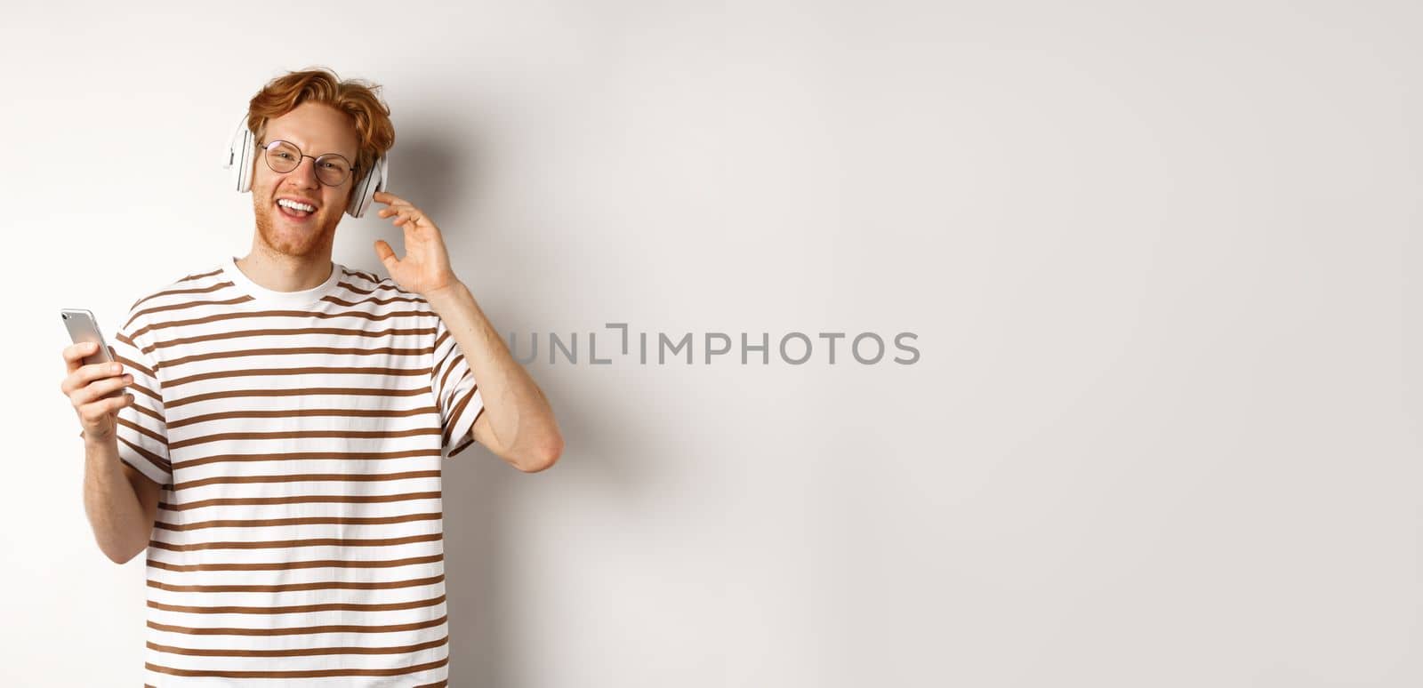 Technology concept. Young man with red hair and beard listening music in headphones and using smartphone, smiling at camera, white background by Benzoix