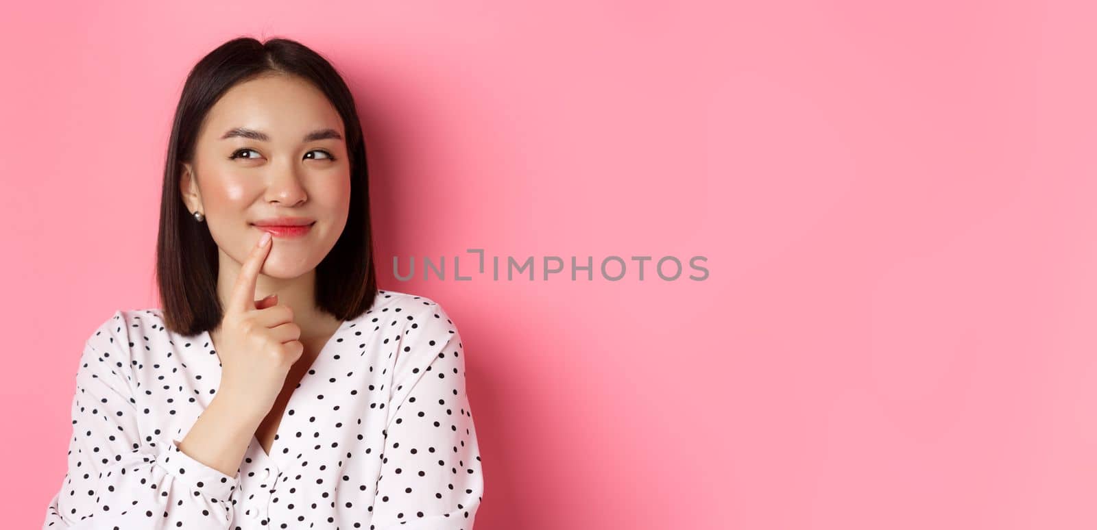 Beauty and lifestyle concept. Close-up of dreamy asian girl smiling, looking left and thinking, making choice, standing over pink background by Benzoix