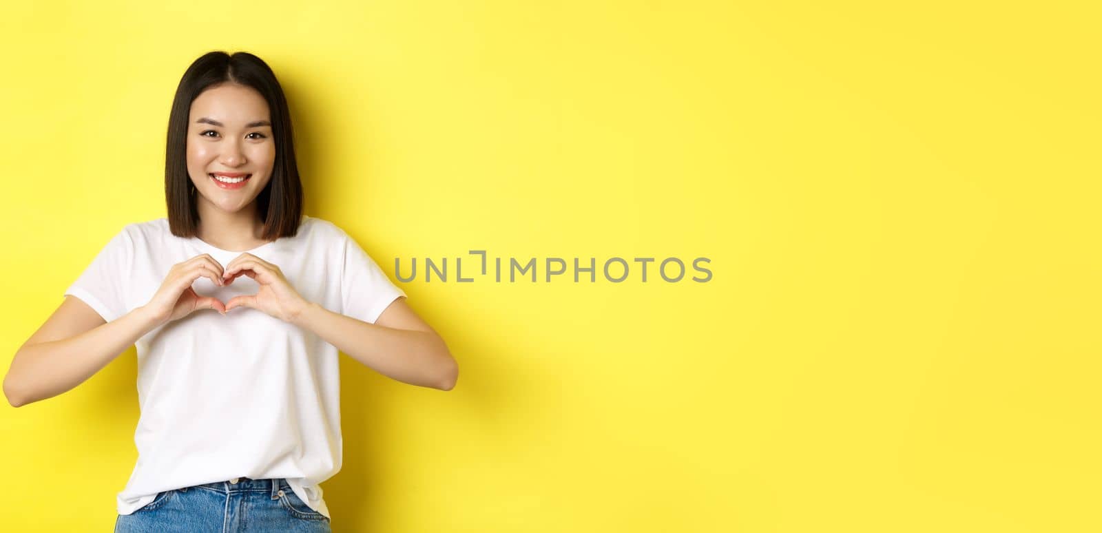 Beautiful asian woman showing I love you heart gesture, smiling at camera, standing against yellow background. Concept of valentines day and romance by Benzoix