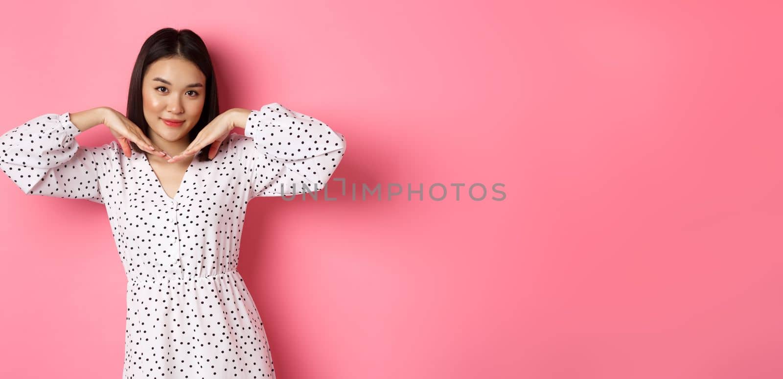 Beautiful asian woman in trendy dress making cute face, holding hands near jawline and gazing coquettish at camera, standing over pink background by Benzoix