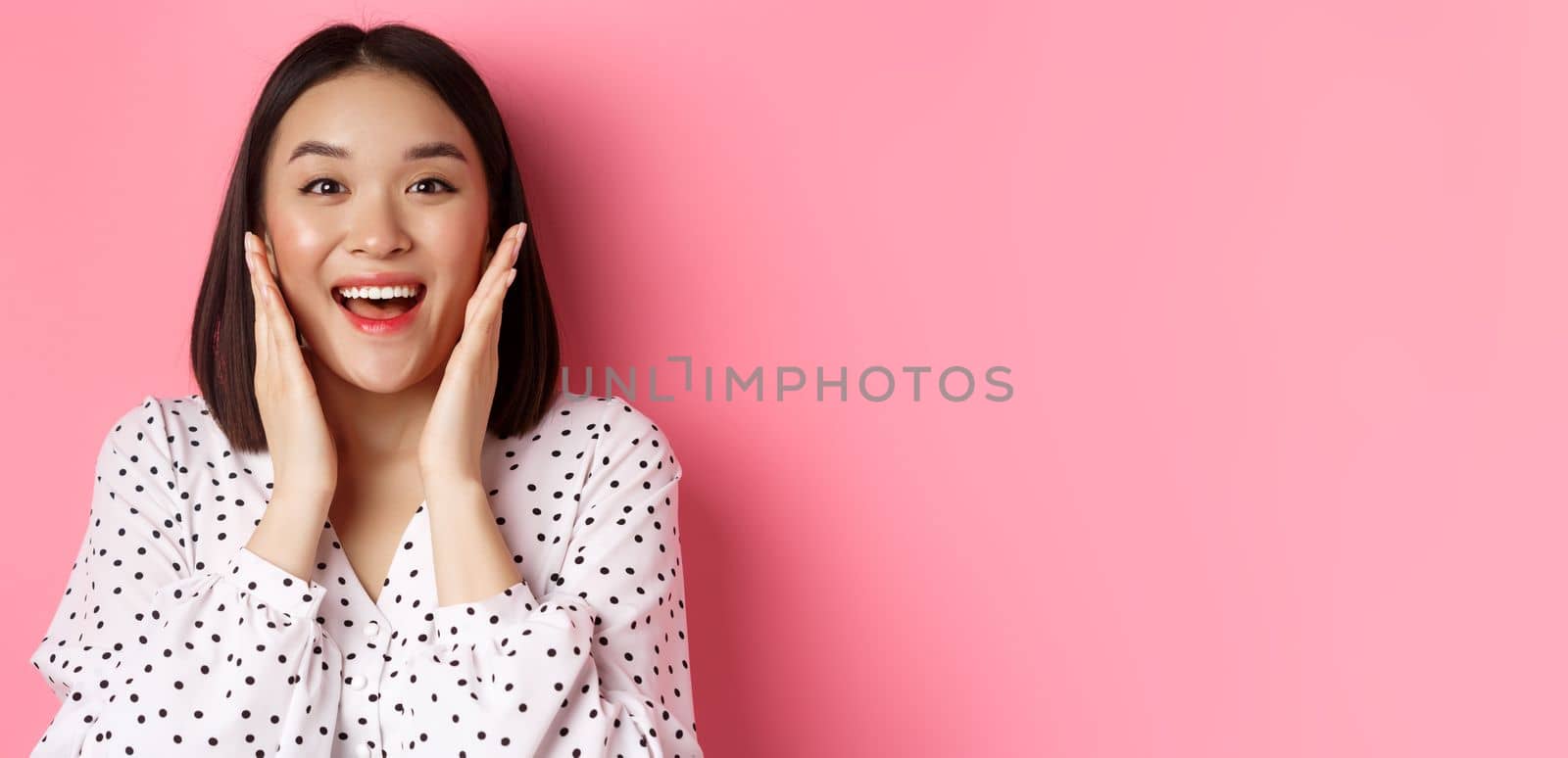 Close-up of beautiful asian woman touching clean skin face and staring at camera amazed, standing over pink background by Benzoix