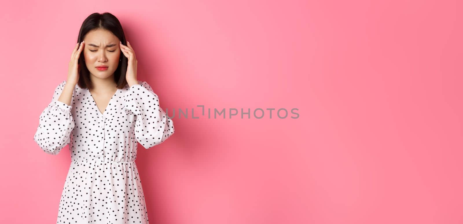 Image of upset asian woman having headache, feeling unwell or dizzy, close eyes and massaging head, suffering migraine, standing over pink background.