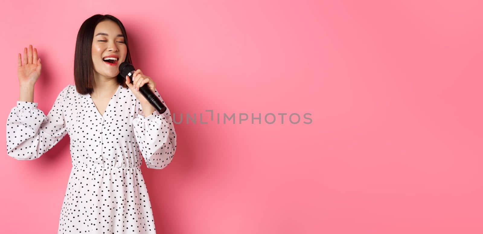 Happy asian woman singing in microphone, having fun at karaoke bar, standing over pink background. Copy space