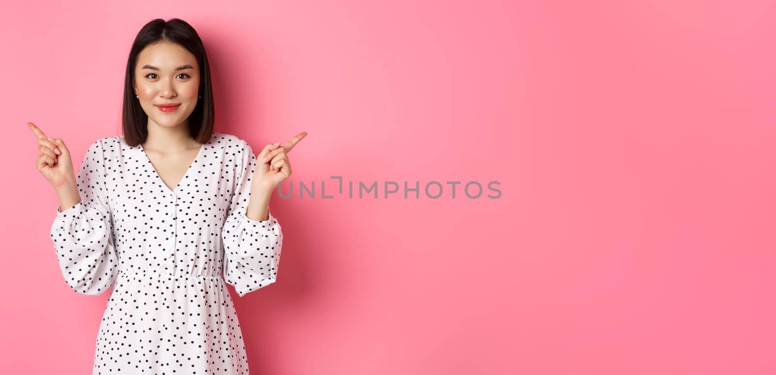 Beautiful asian woman making choice on shopping, pointing fingers sideways and showing variants, smiling at camera, standing over pink background.