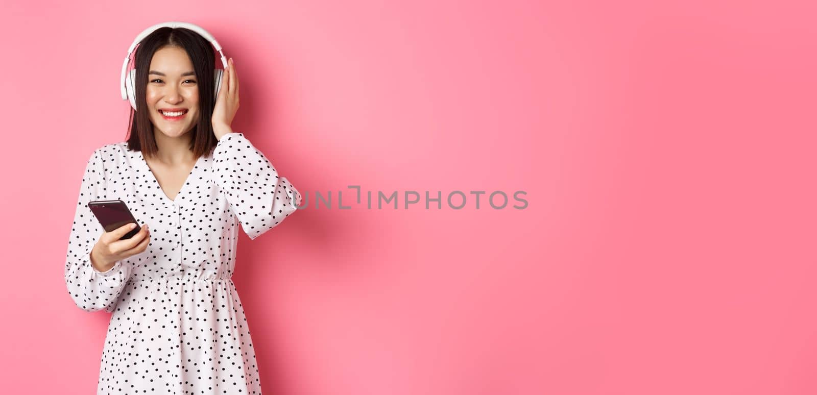 Beautiful asian woman listening music in headphones, using smartphone streaming app and smiling, standing over pink background by Benzoix