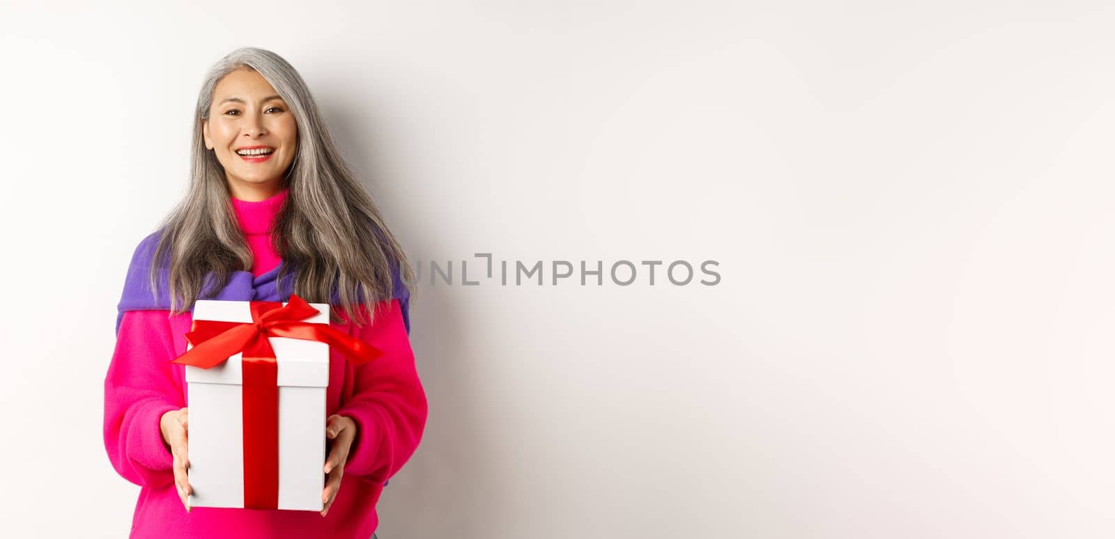 Beautiful asian senior woman smiling, congratulating with valentines day, holding gift in box, standing over white background by Benzoix