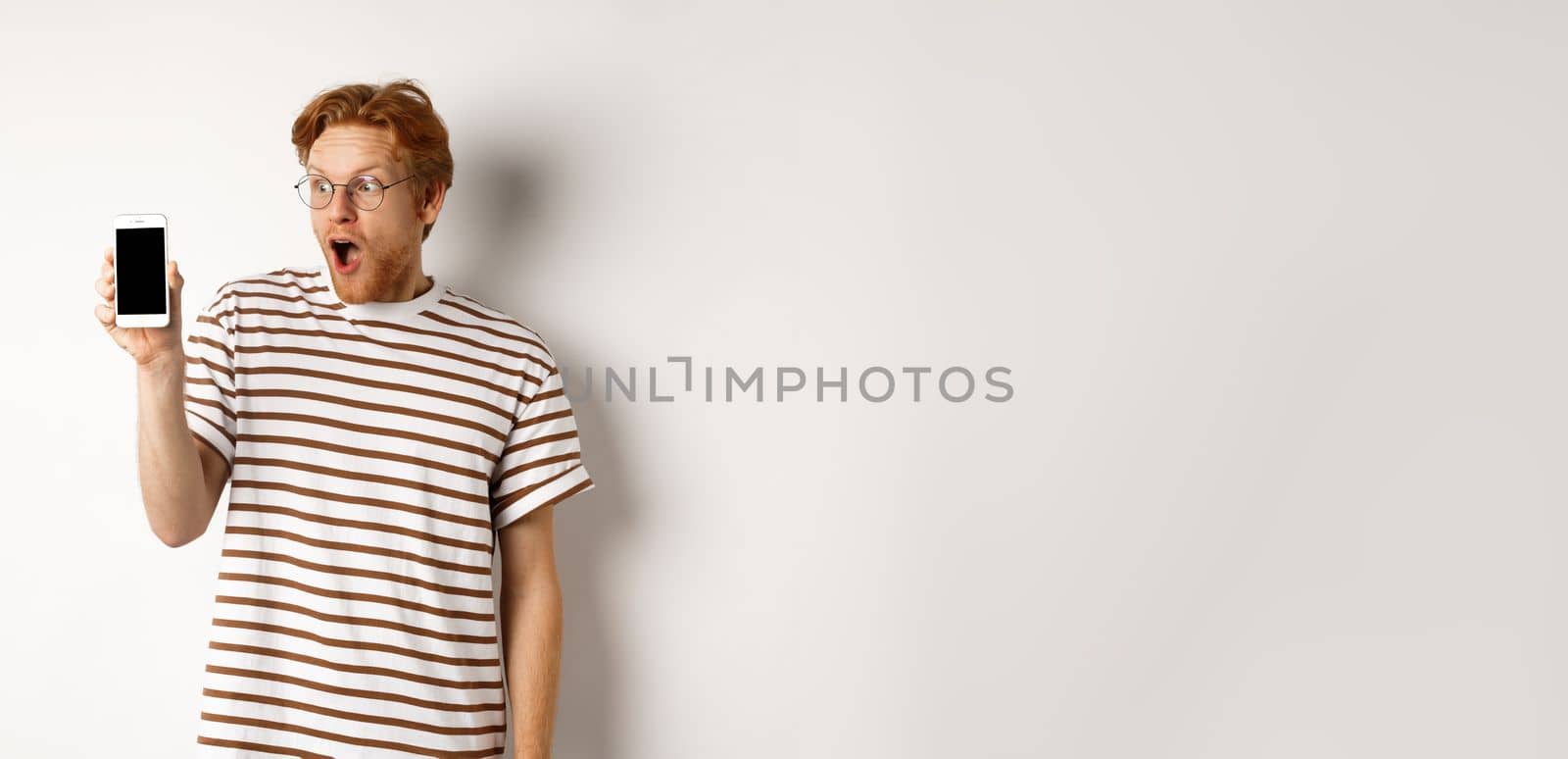 Technology and e-commerce concept. Surprised and shocked redhead guy checking out online promotion, showing blank smartphone screen and drop jaw, white background by Benzoix