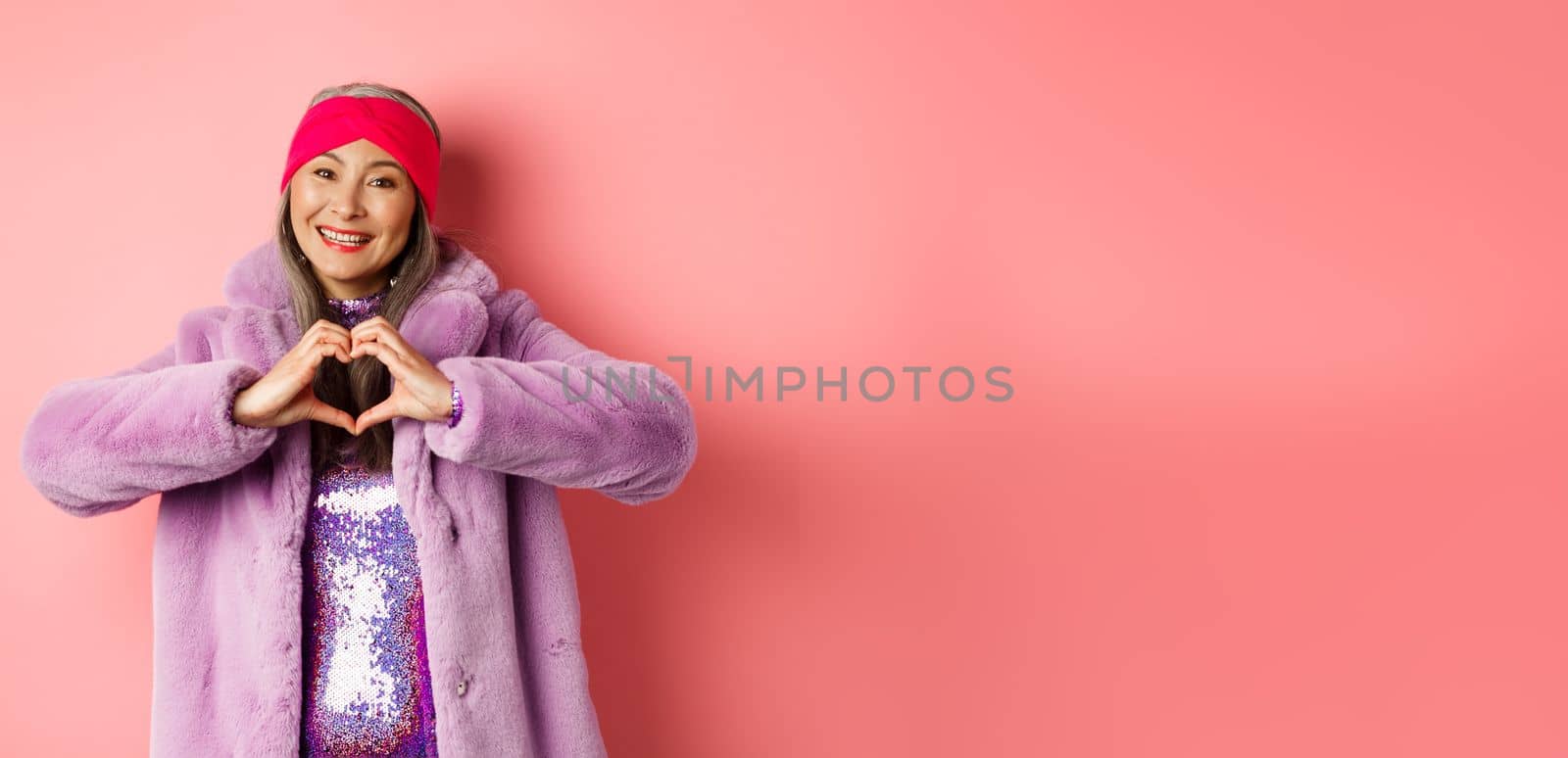 Romance and valentines day. Happy asian senior woman showing heart sign, I love you gesture, smiling and looking caring at camera, standing in faux fur coat, pink background.