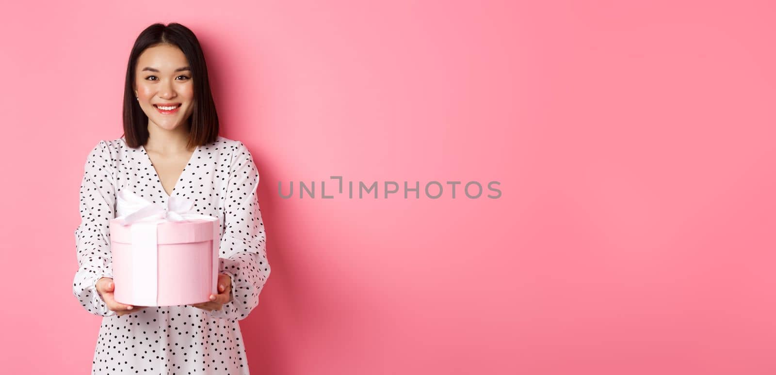 Beautiful asian woman wishing happy holidays, giving you gift in cute box, standing against pink background by Benzoix