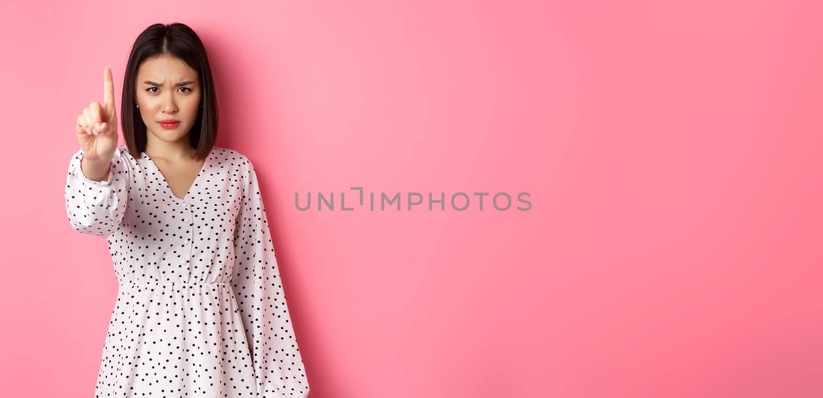 Serious and angry asian woman telling to stop, frowning and showing finger in disapproval, prohibit something, standing in dress against pink background by Benzoix