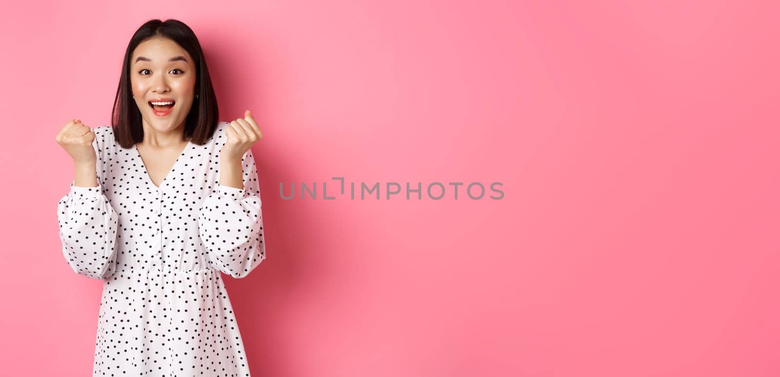 Image of beautiful asian woman winning, clench fists and saying yes with happy face, triumphing and celebrating victory, standing over pink background by Benzoix