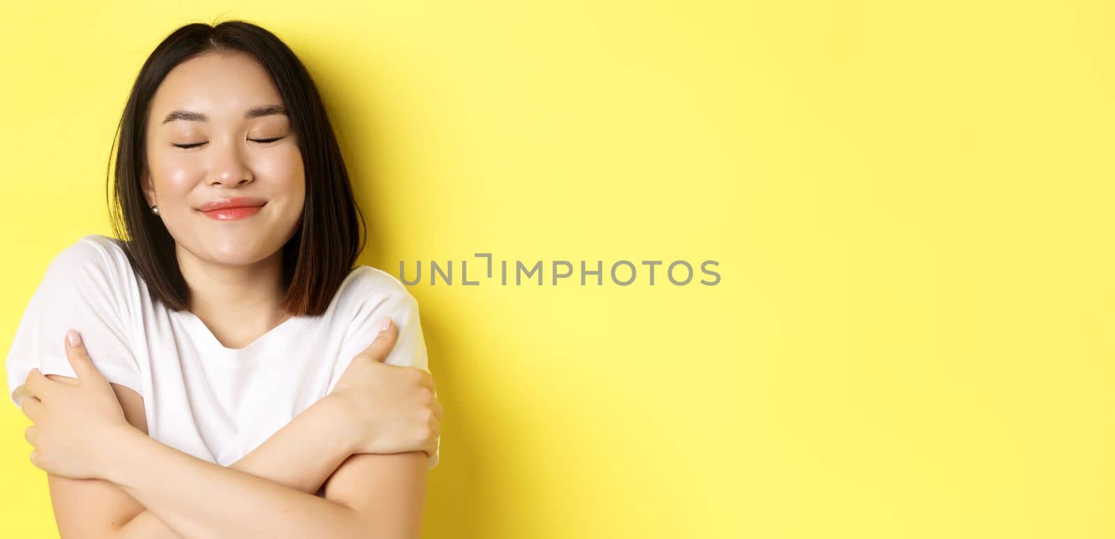 Close up of romantic asian girl hugging herself and dreaming, close eyes and smile while imaging something tender, standing over yellow background.