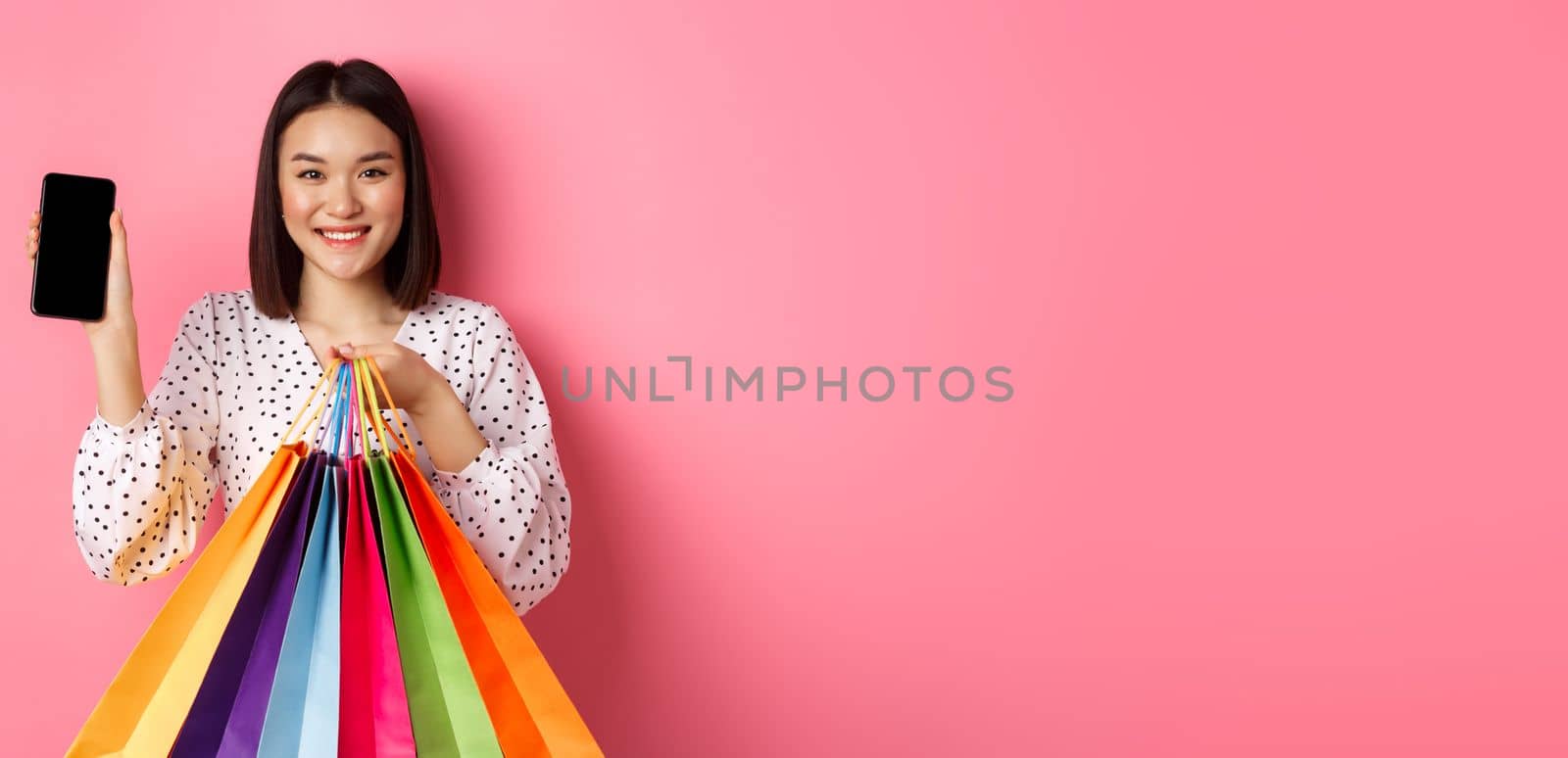 Attractive asian woman showing smartphone app and shopping bags, buying online via application, standing over pink background.