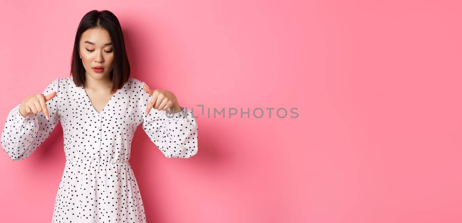 Romantic asian woman peeking down, pointing fingers at bottom, looking curious at product discount, standing over pink background.