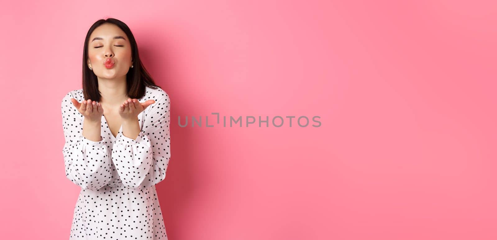 Beautiful chinese girl in dress blowing air kisses, sending mwah at camera, standing over pink background by Benzoix