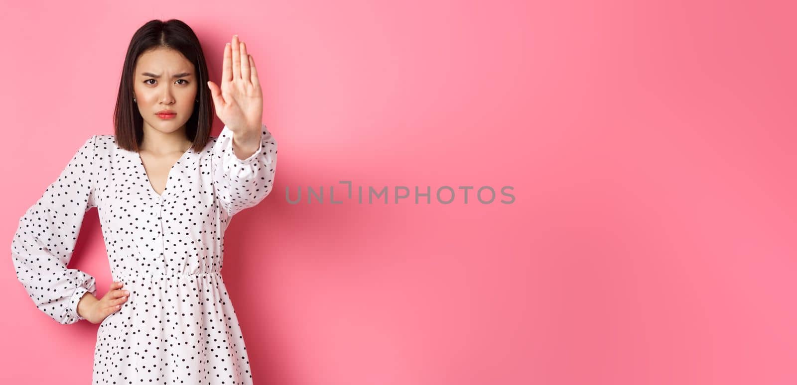 Angry asian woman tell stop, extend arm to prohibit or disapprove something, frowning displeased, standing over pink background by Benzoix