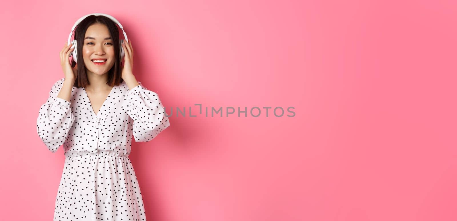 Romantic asian woman smiling happy, listening music in headphones and looking at camera, standing over pink background by Benzoix