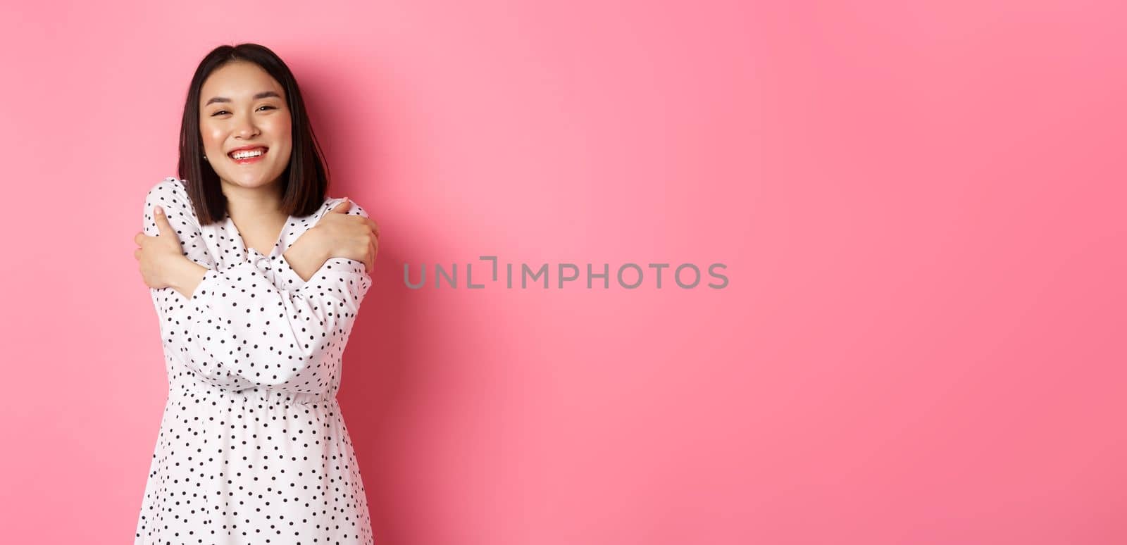 Beautiful asian girl embracing own body, hugging herself and smiling with happy face, looking at camera carefree, standing over pink background.